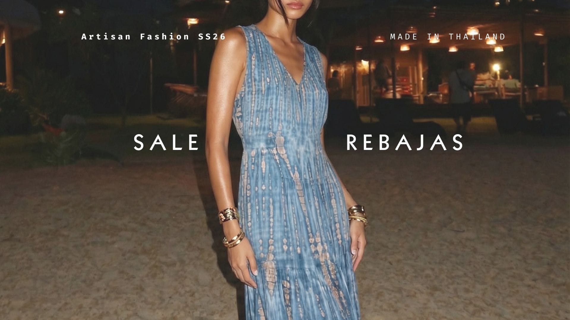 A woman wearing a sleeveless blue patterned maxi dress stands outdoors at night in front of a softly lit venue. White text overlays read 'Artisan Fashion SS26', 'MADE IN THAILAND', 'SALE', and 'REBAJAS'.